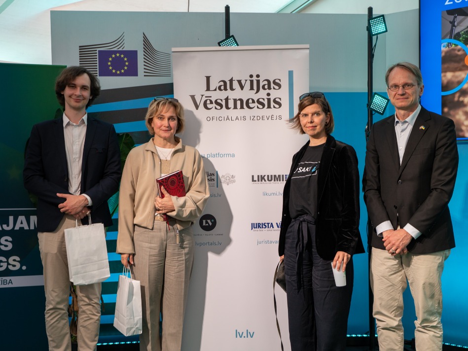 Četri diskusijas "Tiesībpratības stafete" dalībnieki pozē pie oficiālā izdevēja "Latvijas Vēstnesis" banera sarunu festivālā "LAMPA".